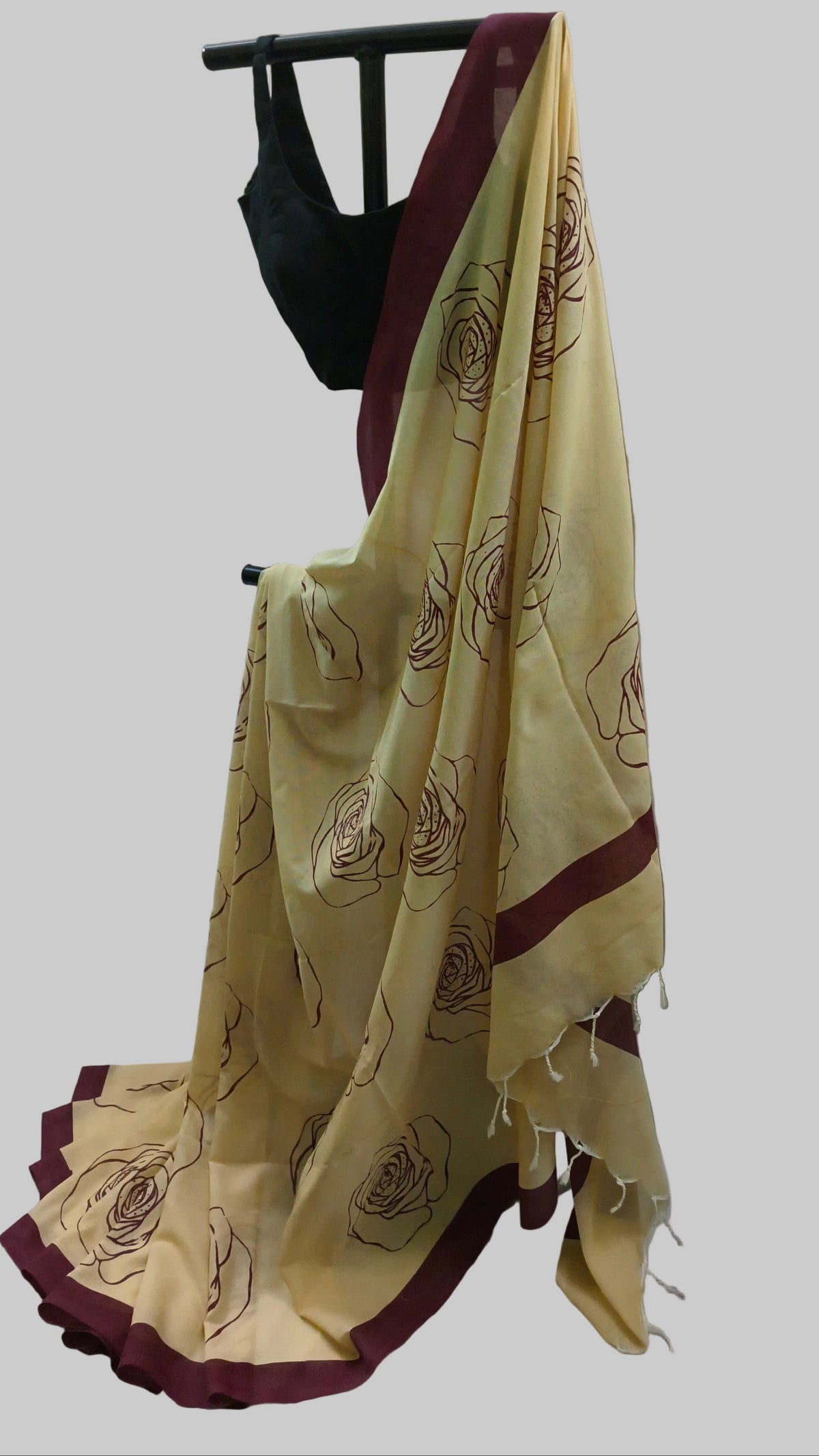 A cream-colored saree with dark brown floral patterns and deep maroon borders, draped on a black clothing stand with a black blouse, set against a neutral beige curtain backdrop.