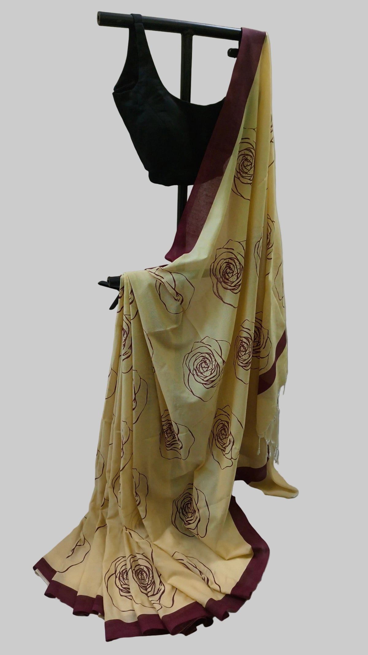 A beige cotton saree with dark maroon borders and brown floral line prints, draped on a black clothing stand alongside a black sleeveless blouse against a cream curtain backdrop.