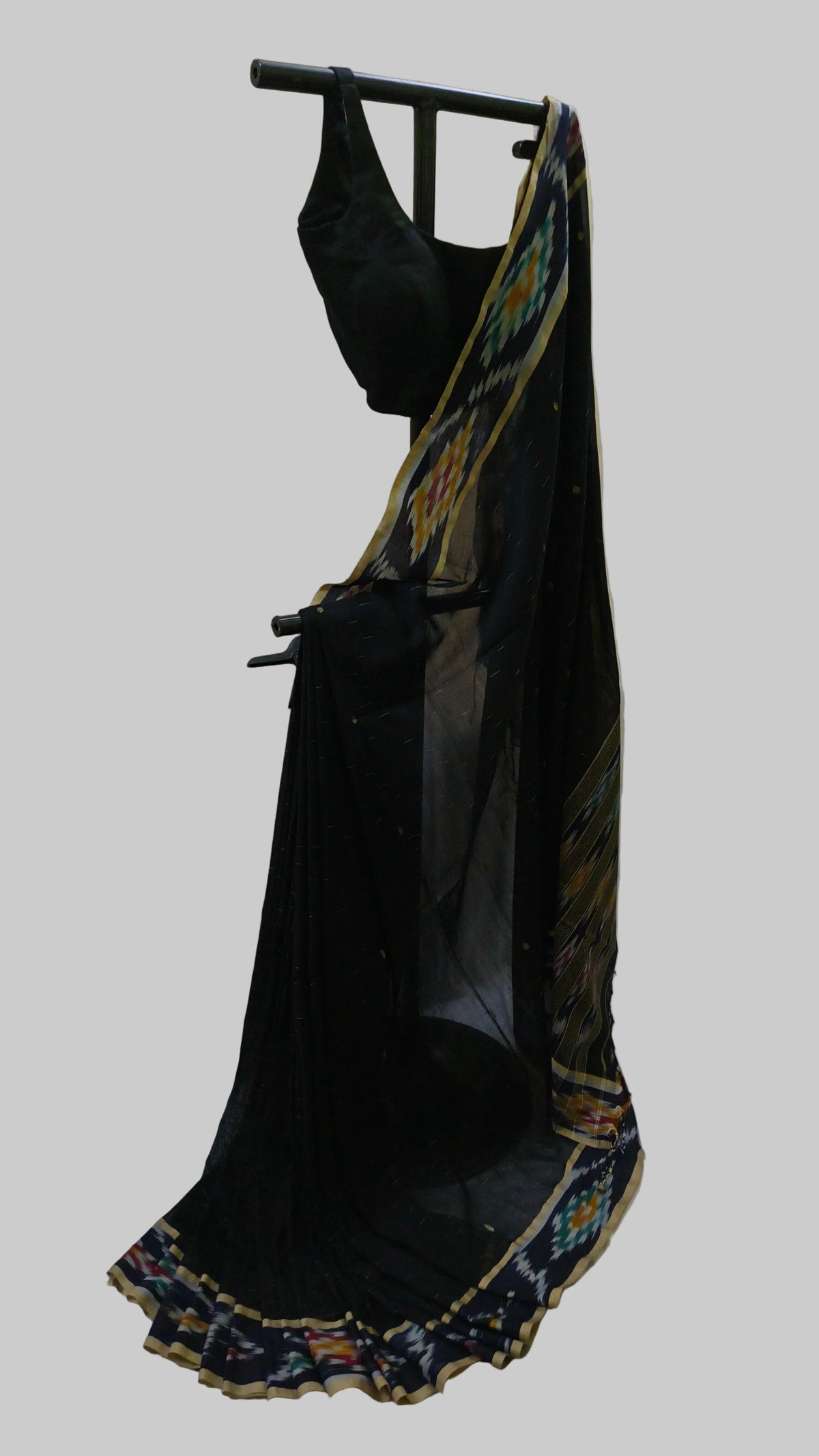 A black georgette saree with a multicolor ikat border and a matching black blouse, displayed on a black clothing rack against a beige curtain backdrop.