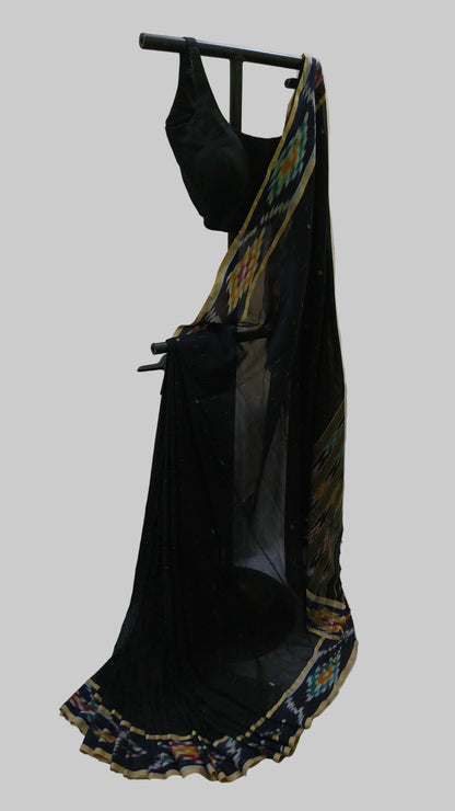 A black georgette saree with a multicolor ikat border and a matching black blouse, displayed on a black clothing rack against a beige curtain backdrop.