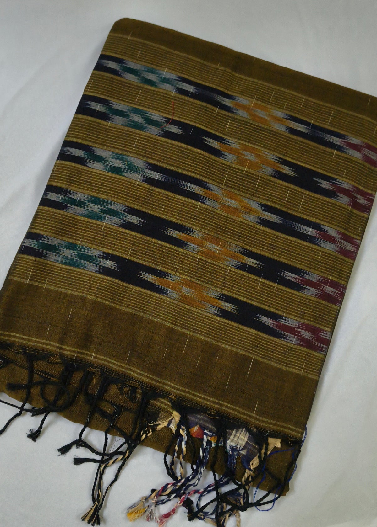 A folded handwoven textile, likely a traditional sari, featuring horizontal stripes with geometric ikat patterns in black, navy, teal, orange, and purple on a golden-brown base, finished with a fringed tassel edge.