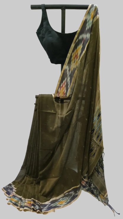 An olive green silk saree draped on a black clothing hanger with a black sleeveless blouse, set against a neutral cream-colored fabric backdrop.