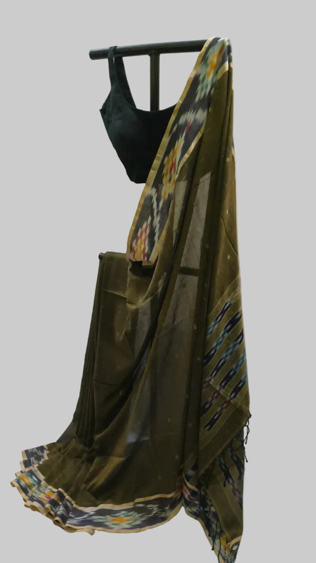 An olive green silk saree draped on a black hanger with a black blouse, set against a cream-colored curtain backdrop.