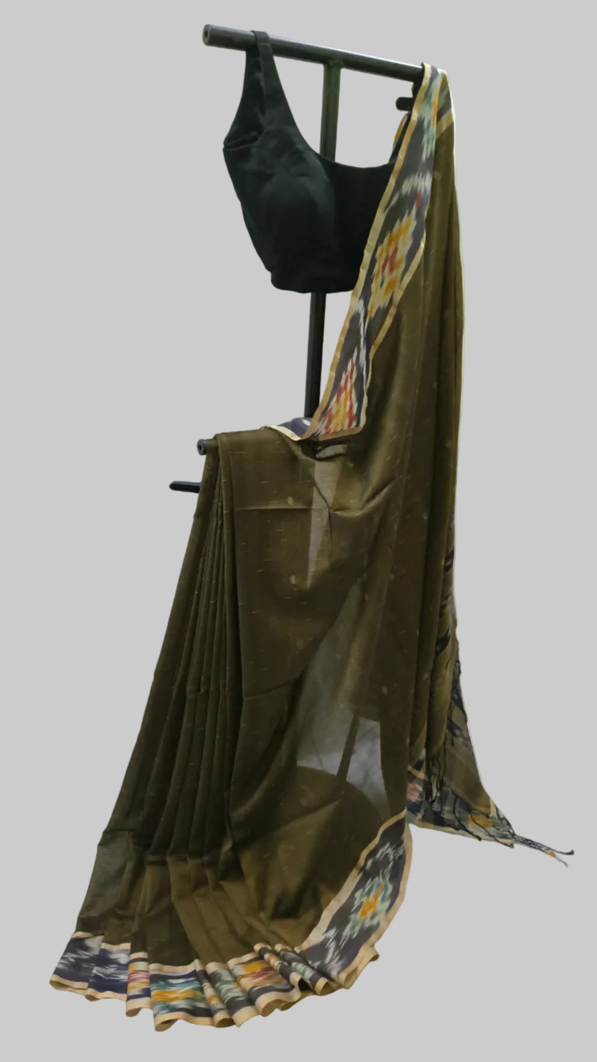 An olive green cotton saree draped on a black clothing stand, paired with a black sleeveless blouse. The saree features a multicolor patterned border and is set against a neutral draped backdrop.