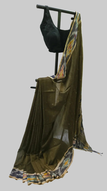 An olive green cotton saree draped on a black clothing stand, paired with a black sleeveless blouse. The saree features a multicolor patterned border and is set against a neutral draped backdrop.