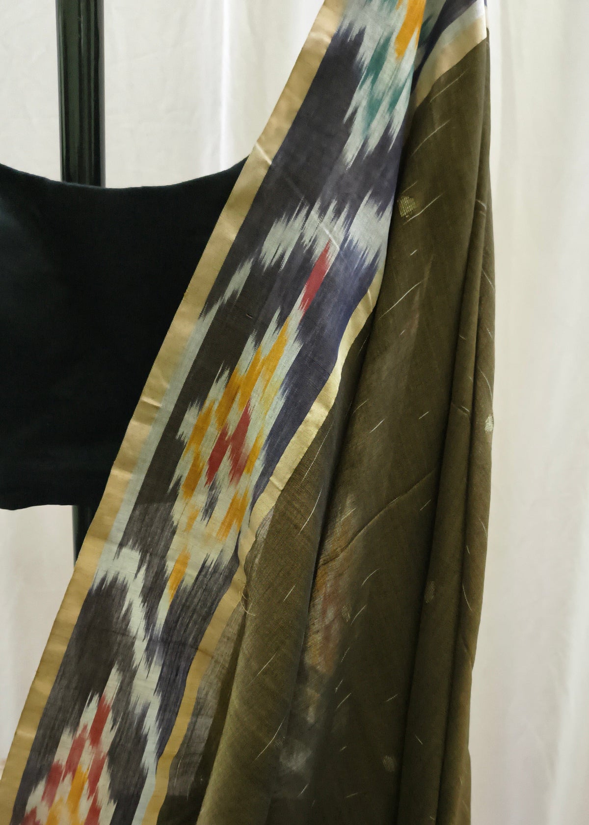 An olive green cotton saree draped over a black mannequin, featuring a wide border with a multicolor ikat pattern in yellow, red, blue, and white, and a gold zari border.
