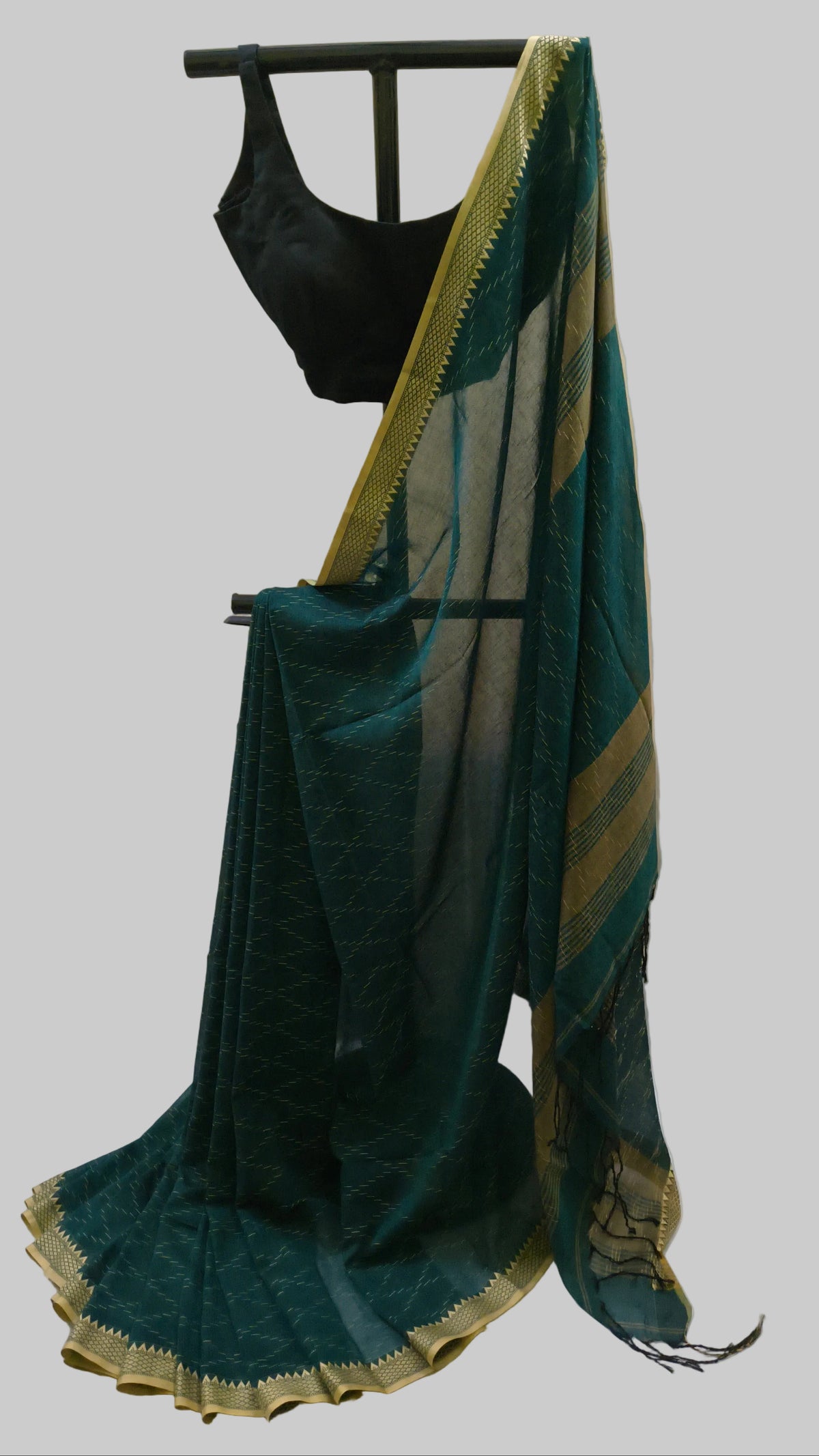 A dark teal green saree with gold zari borders and a black blouse, displayed on a black clothing rack against a beige curtain backdrop.