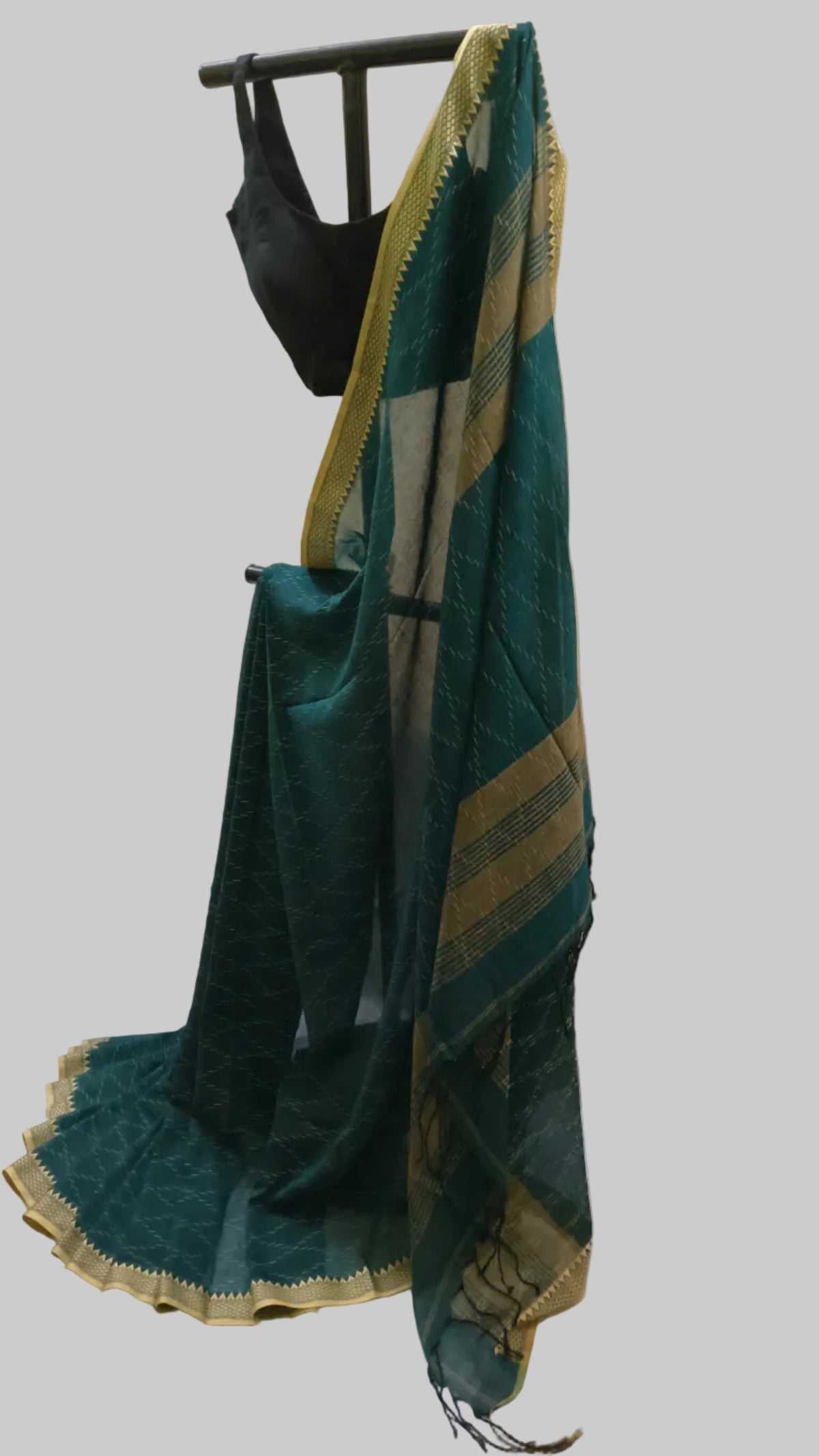 A dark teal green saree with gold zari border detailing draped over a black hanger, paired with a black blouse, set against a cream-colored curtain backdrop.