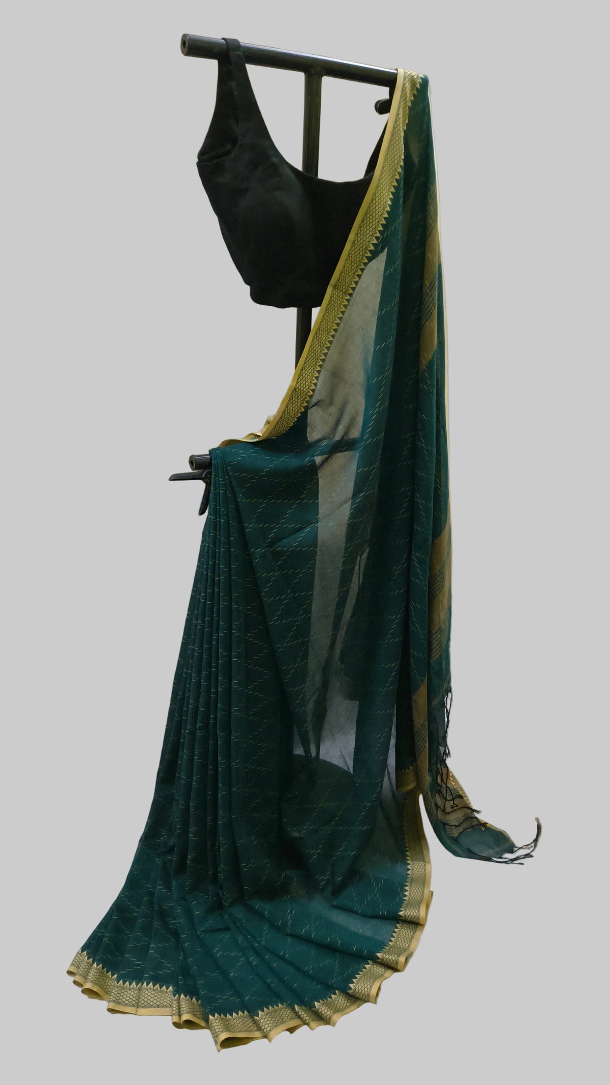 A dark teal green saree with a gold border, draped on a black clothing stand alongside a black blouse, set against a cream-colored curtain backdrop.
