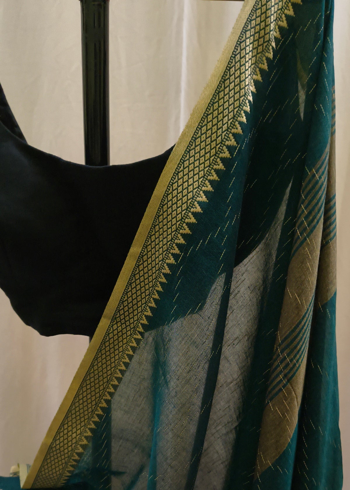 A close-up of a dark teal-green saree or fabric drape with a wide gold border and subtle woven pattern, shown hanging against a light neutral backdrop with a black blouse partially visible.