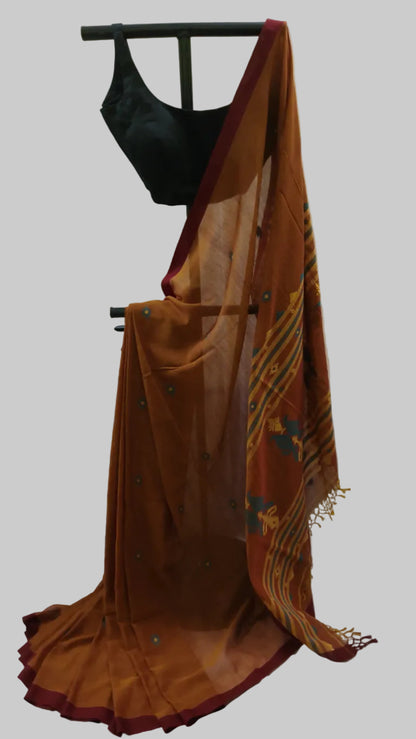 A rust-orange saree with a deep maroon border and yellow striped motifs, draped on a black dress form with a black sleeveless blouse, set against a cream-colored curtain backdrop.