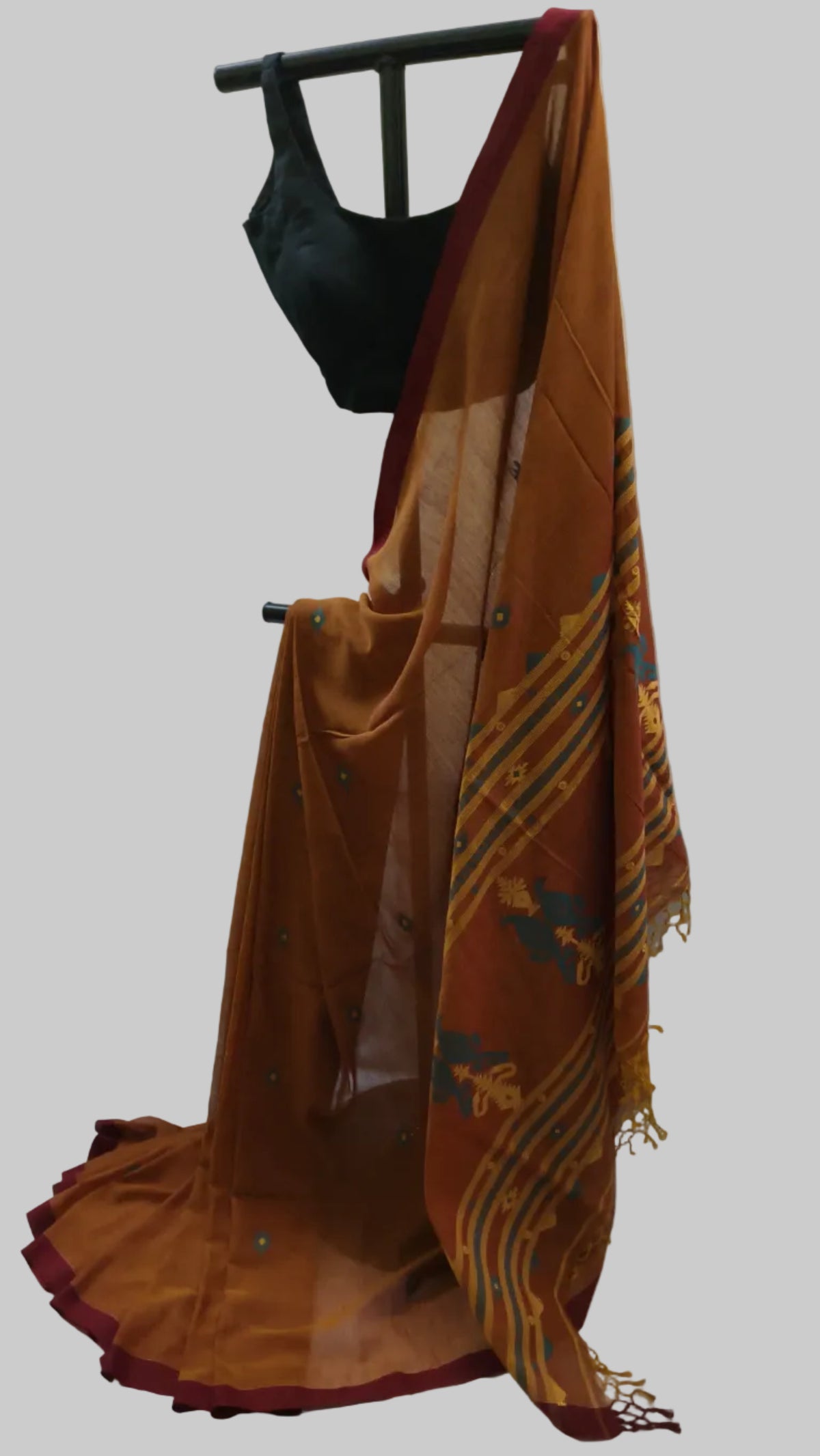 An orange-brown sheer saree with yellow and blue woven patterns and a dark red border, draped on a black clothing stand with a black blouse against a cream fabric backdrop.