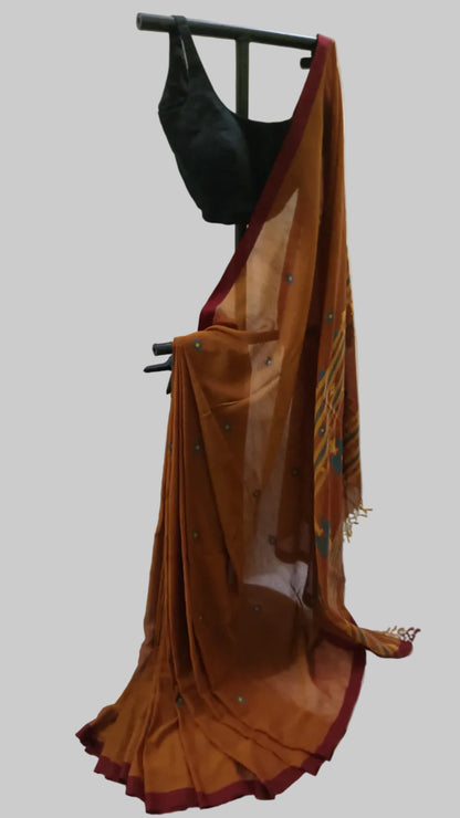 A rust-orange saree with a deep maroon border and small embroidered motifs, paired with a black sleeveless blouse, displayed on a black hanger against a draped cream backdrop.