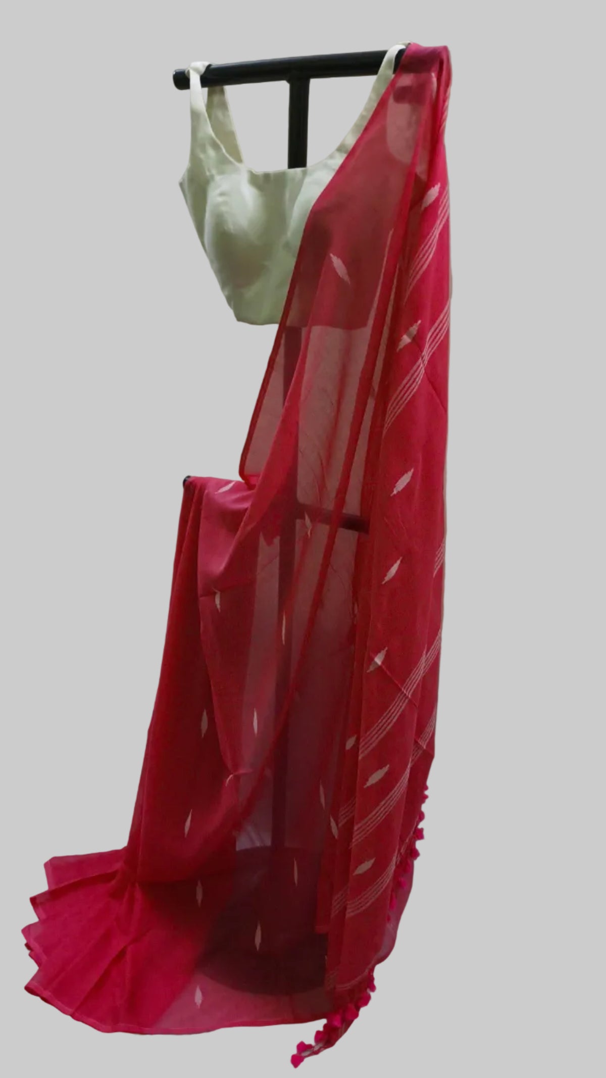 A bright pink-red saree with white geometric patterns and a matching light green blouse, displayed on a black hanger against a draped white fabric backdrop.
