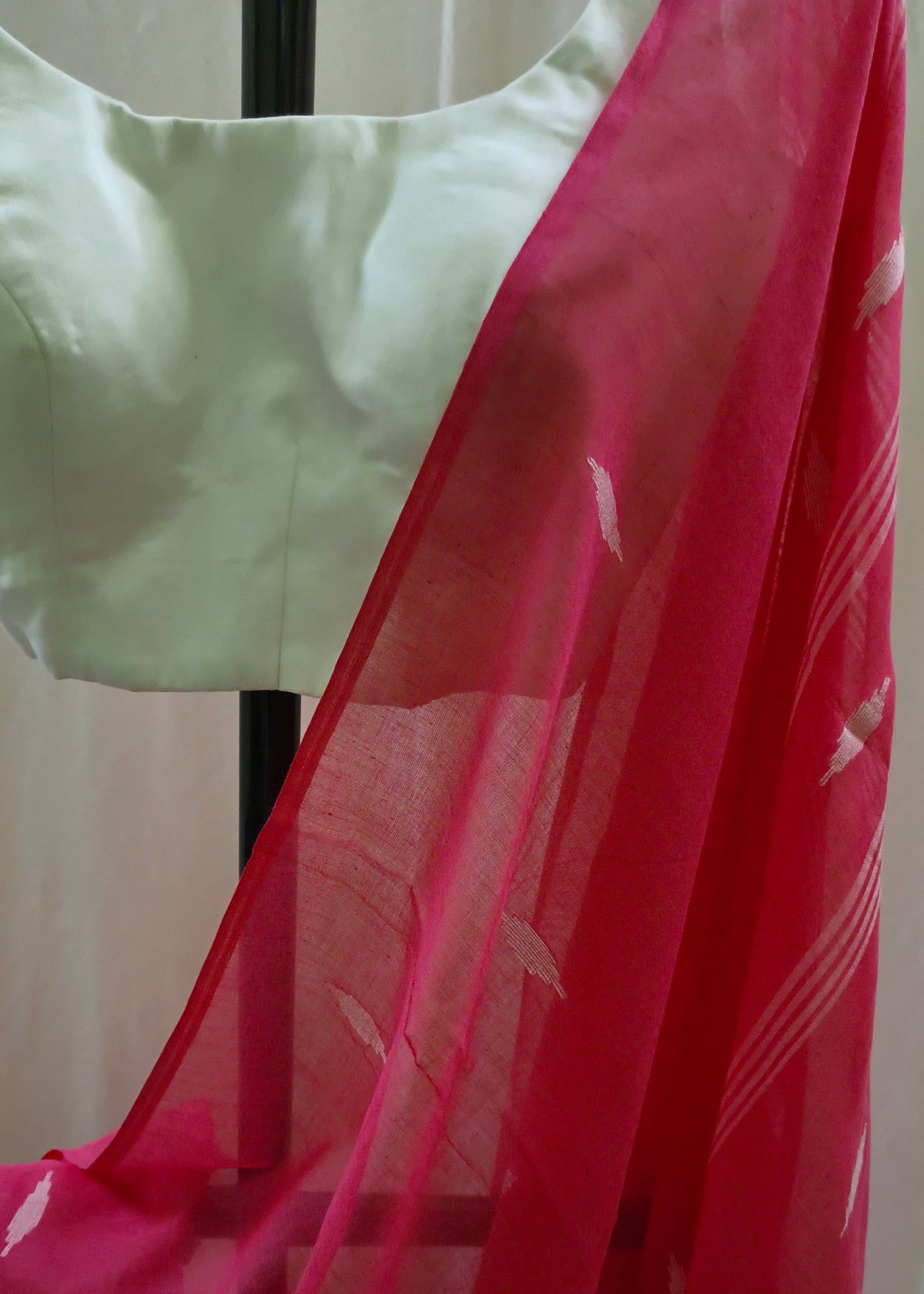 A close-up of a bright pink sheer saree drape with small white printed motifs, paired with a light cream blouse on a black hanger.