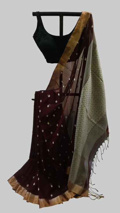 A traditional Indian saree displayed on a black hanger against a neutral draped backdrop. The saree features a deep maroon body with small gold motifs, a beige patterned border, and a metallic gold zari border, paired with a black blouse.