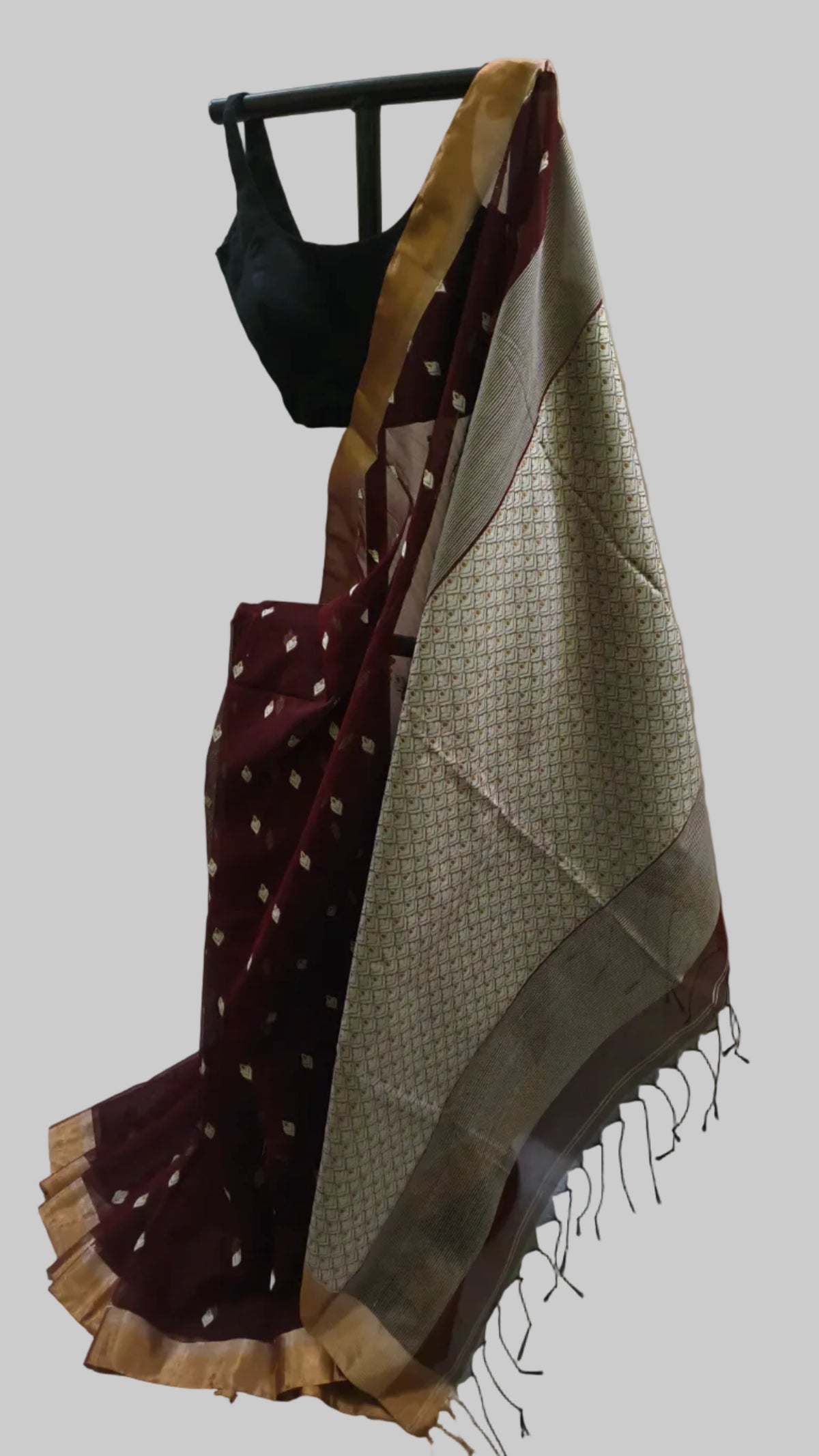 A traditional Indian saree draped on a black hanger stand, featuring a deep maroon body with small gold motifs, a beige patterned pallu, and a gold border, paired with a black blouse.