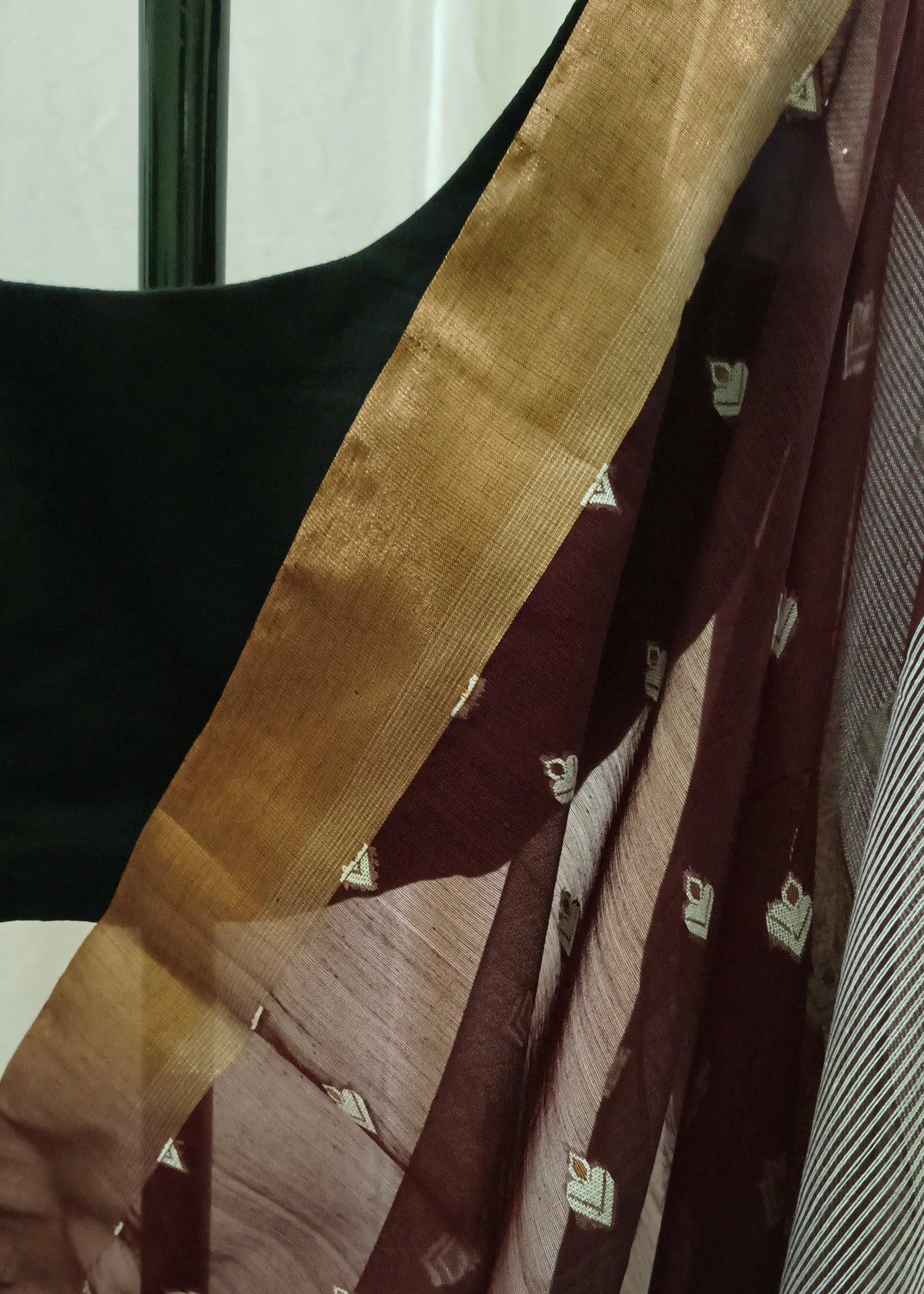 A close-up of a draped saree or fabric panel with a wide metallic gold border, a deep maroon body, and small white motifs woven or printed throughout.