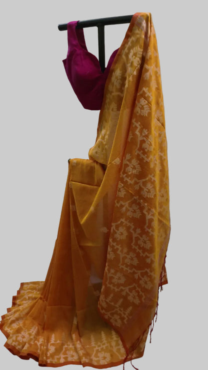 A yellow-orange saree with a white floral pattern draped over a black hanger, paired with a magenta pink sleeveless blouse, set against a white curtain backdrop.
