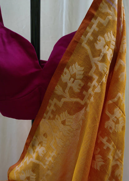 A close-up of a yellow-orange saree with a beige floral patterned border, draped over a magenta blouse against a white curtain background.