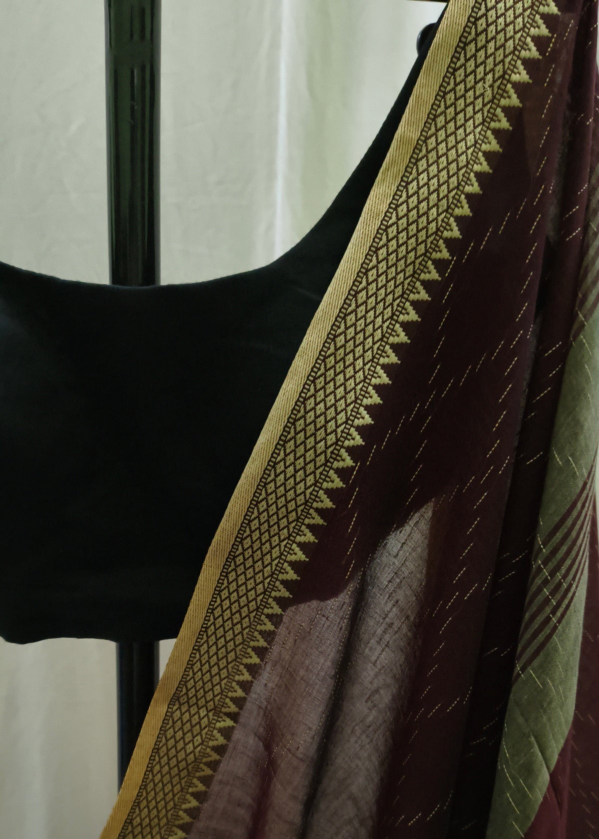 A close-up of a draped saree or fabric panel in deep maroon with a wide gold patterned border, shown on a black dress form against a light neutral background.