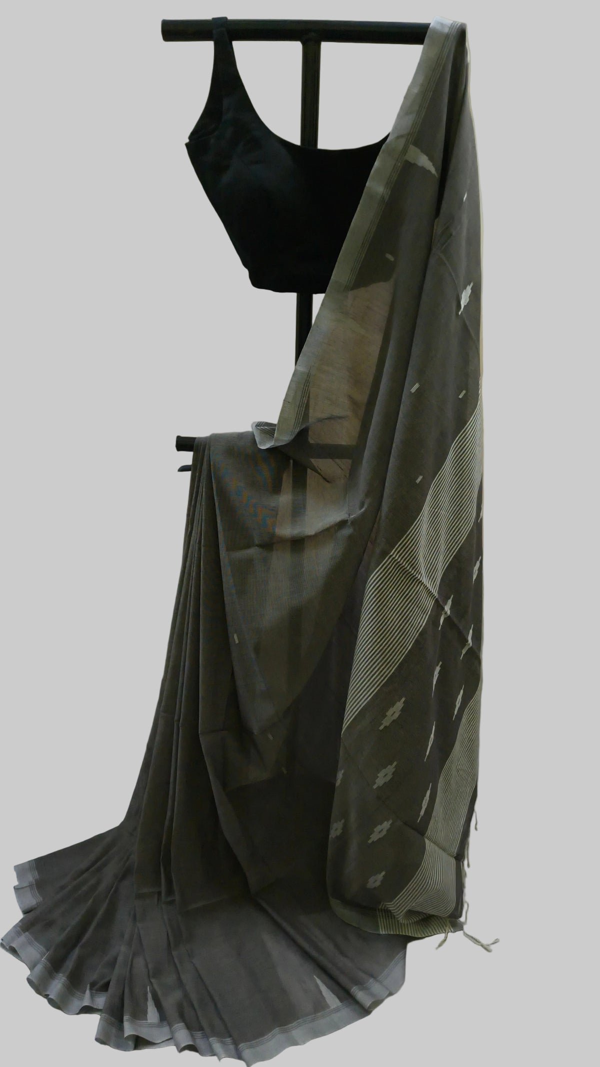 A dark gray saree with a subtle pattern and a black sleeveless blouse, displayed on a black clothing stand against a beige curtain backdrop.