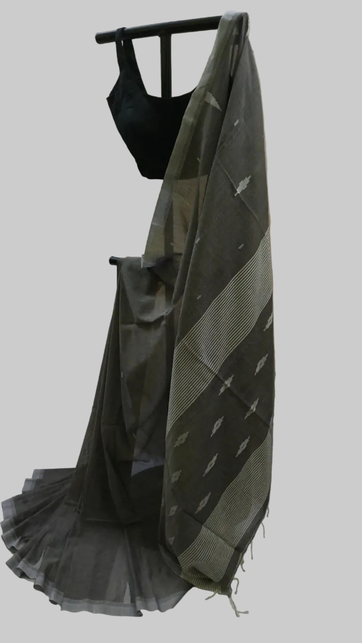 A dark gray and beige patterned saree draped on a black dress form, paired with a black sleeveless blouse, set against a neutral cream-colored fabric backdrop.
