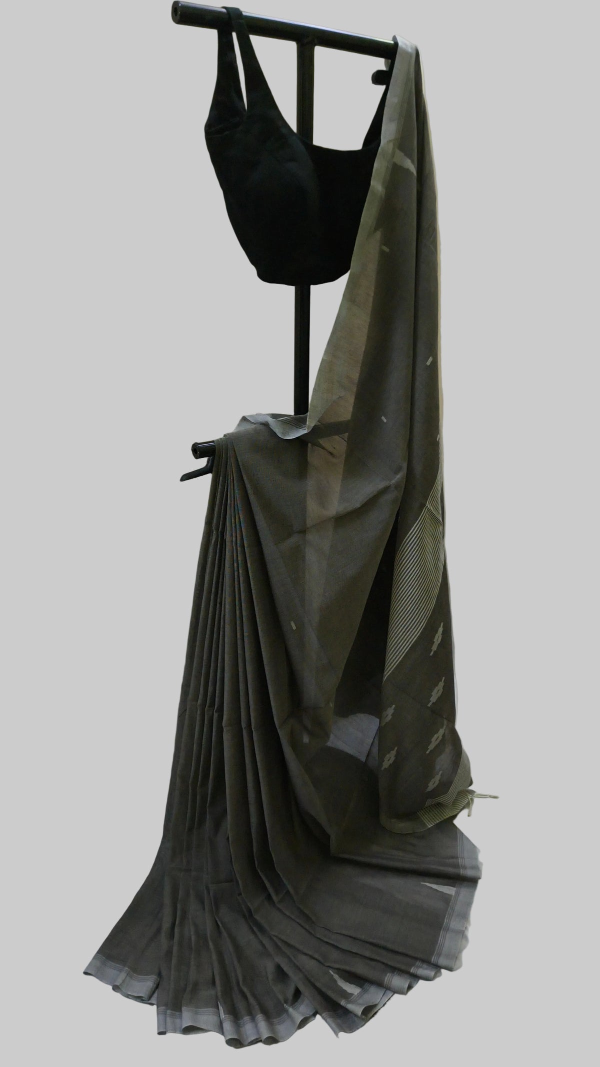 A dark gray pleated saree with a black blouse displayed on a black dress form, set against a draped beige fabric backdrop.
