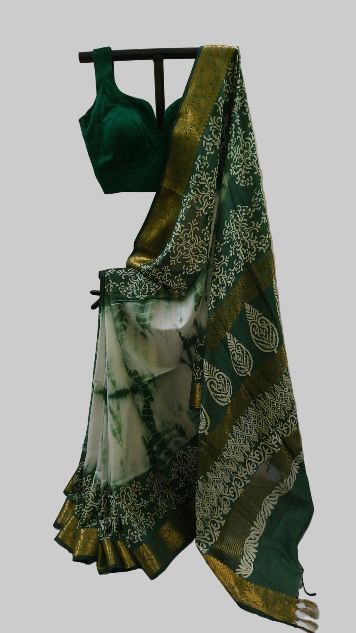 A dark green saree with a white and green tie-dye patterned body, a gold zari border, and a matching dark green blouse, displayed on a stand against a beige curtain backdrop.
