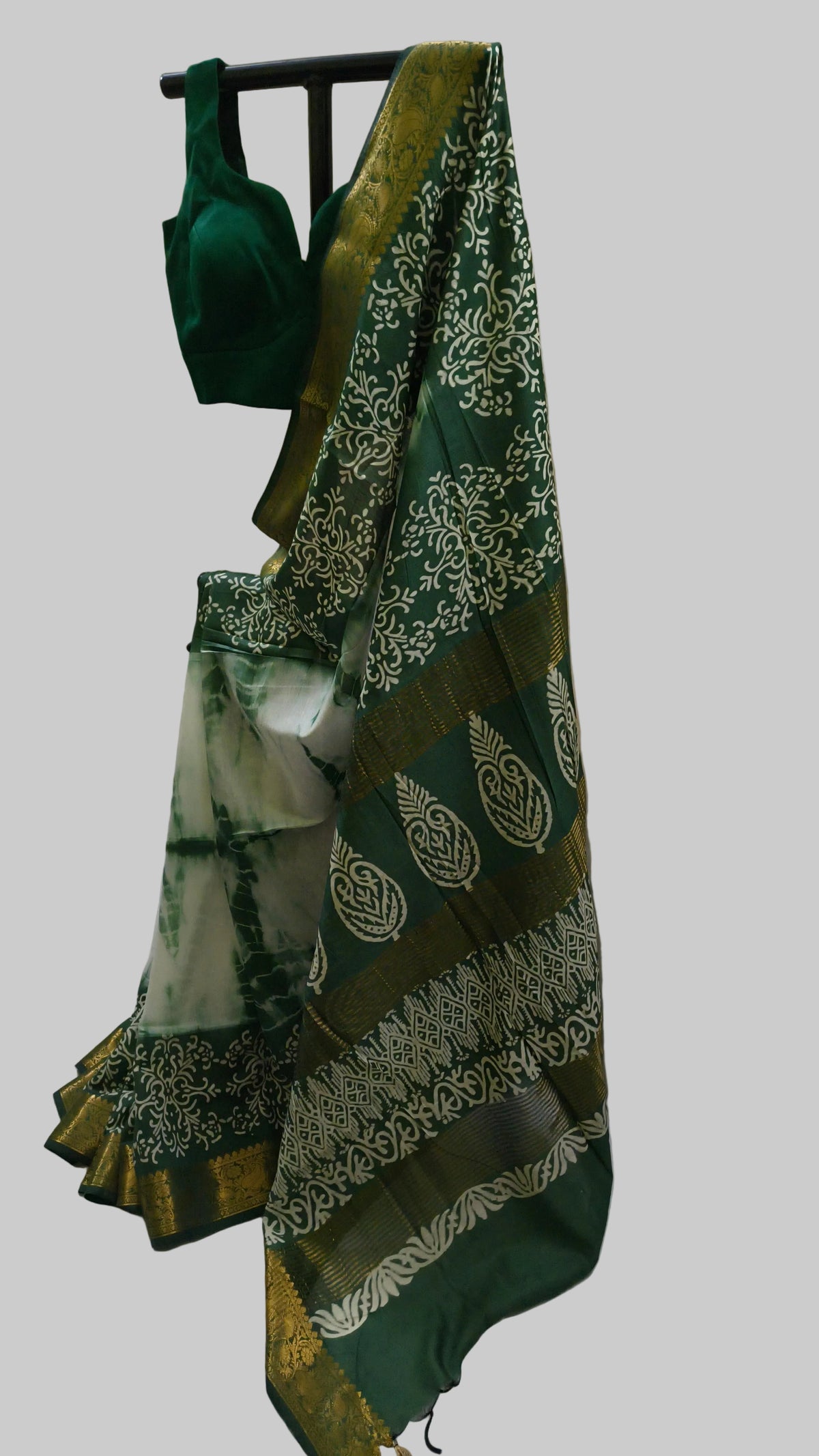 A dark green saree with white floral patterns and a gold border, paired with a matching dark green blouse, displayed on a stand against a cream-colored curtain backdrop.