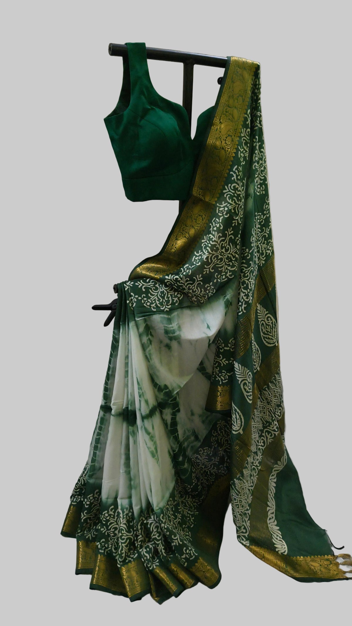 A dark green and white tie-dye saree with a wide gold zari border and matching green blouse displayed on a black hanger against a cream curtain backdrop.