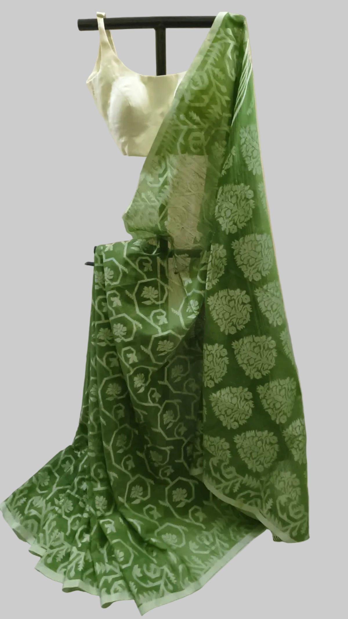 A green saree with a light green floral and vine pattern, draped on a black hanger with a light green blouse, set against a neutral beige curtain background.