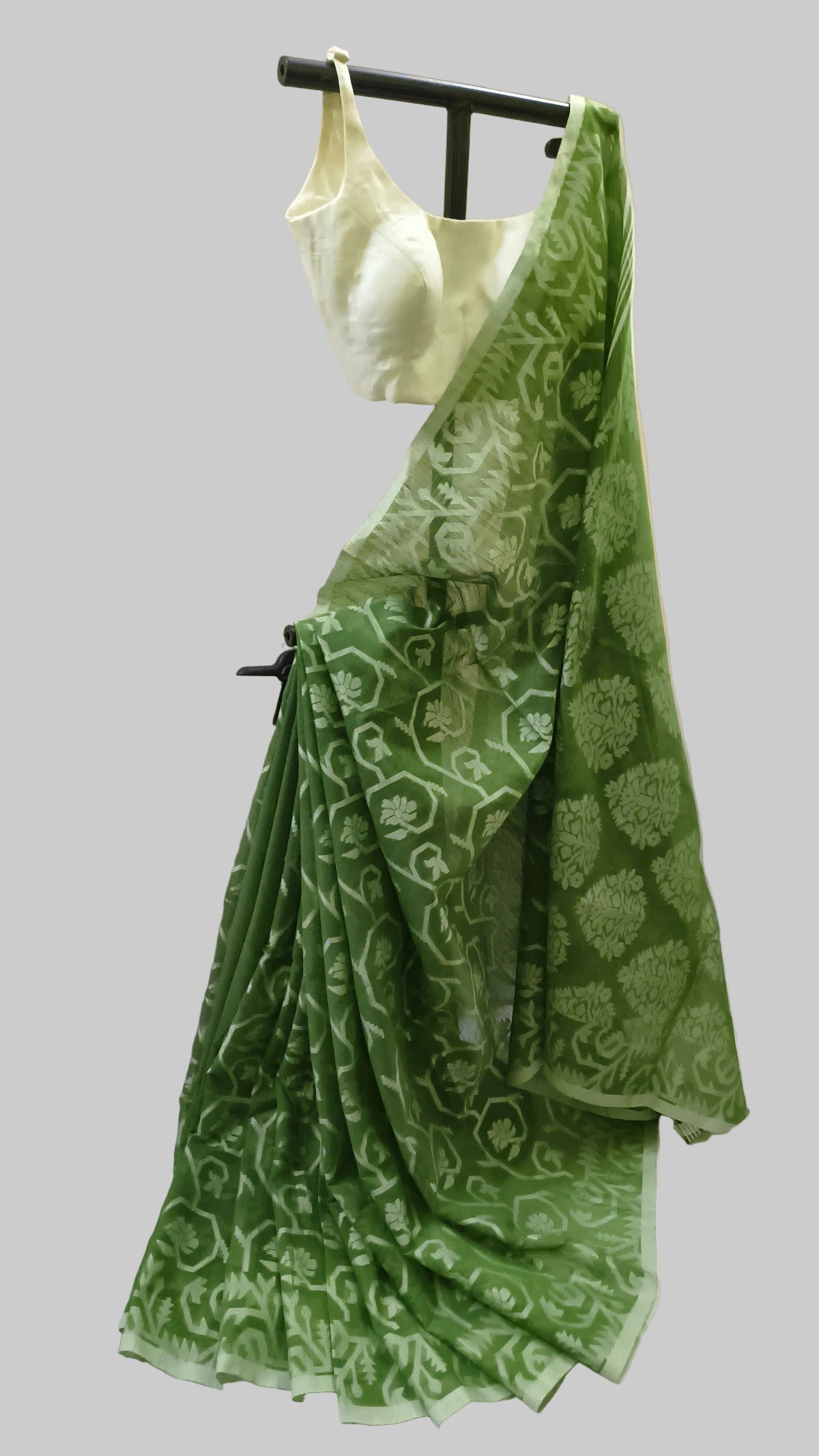 A green patterned saree with a matching light green blouse displayed on a black hanger against a beige curtain backdrop.