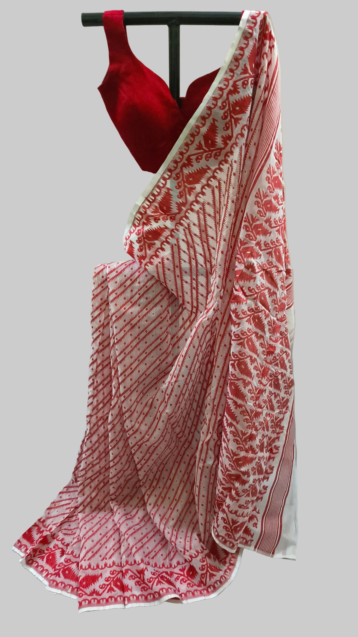 A red and white patterned saree draped on a black hanger with a matching red blouse, set against a neutral cream-colored backdrop.