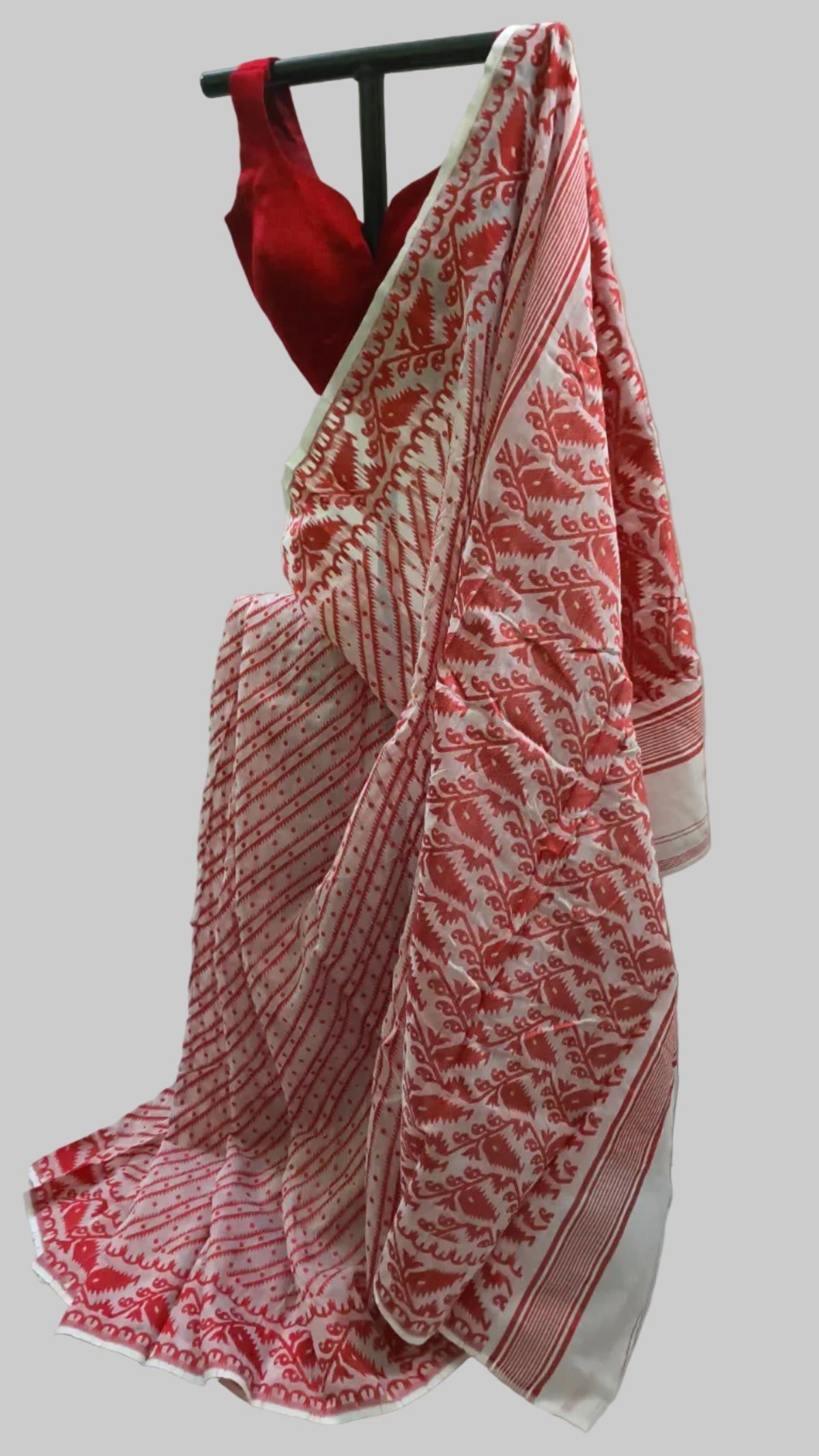 A red and white patterned saree draped on a black hanger with a red sleeveless blouse, set against a white fabric backdrop.