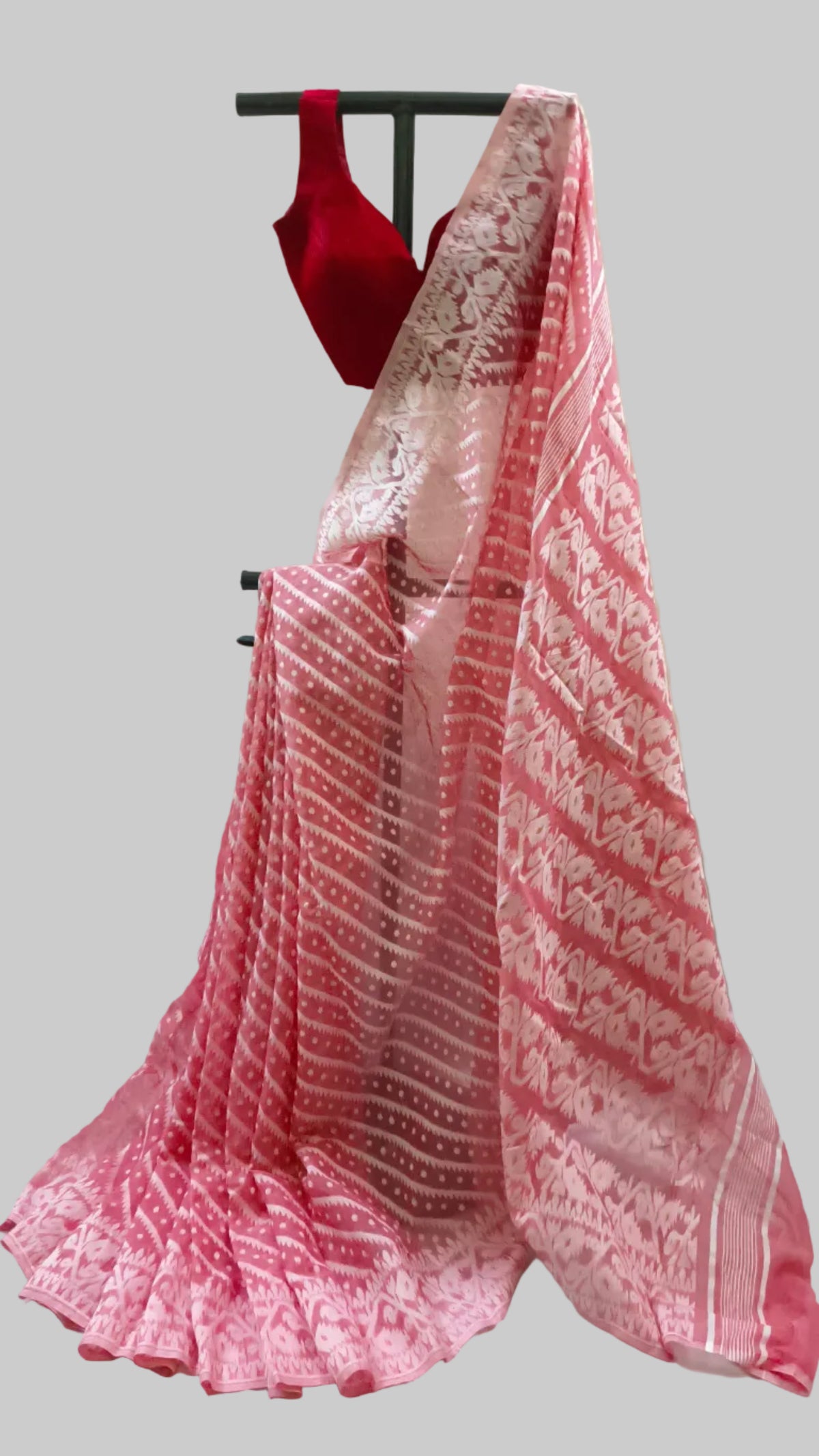 A pink and red patterned saree draped on a black hanger stand, paired with a red sleeveless blouse, set against a draped cream fabric backdrop.