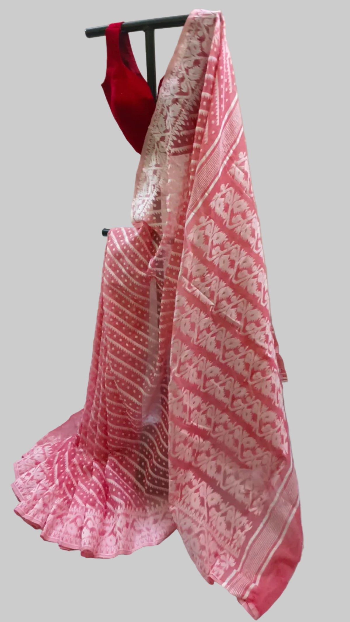 A pink and white patterned saree draped on a black hanger stand, paired with a red sleeveless blouse, set against a draped cream fabric backdrop.