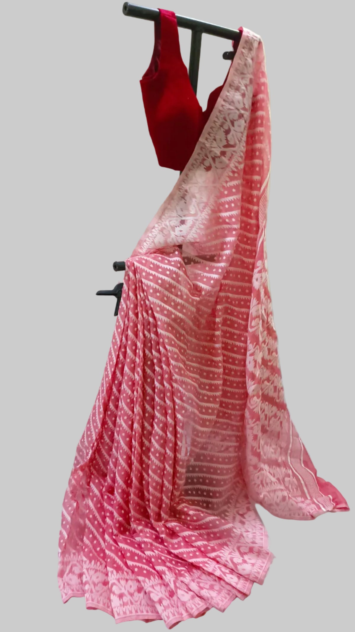 A pink and red patterned saree draped on a black clothing stand, paired with a solid red sleeveless blouse, set against a white fabric backdrop.