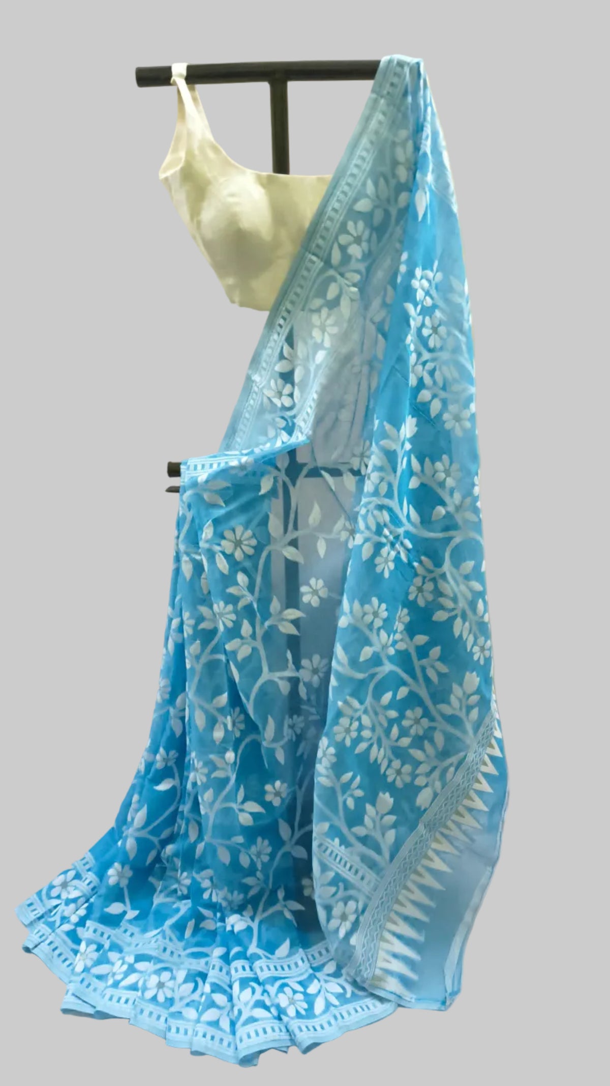 A light blue embroidered saree with white floral patterns and a matching blouse displayed on a black hanger against a beige fabric backdrop.