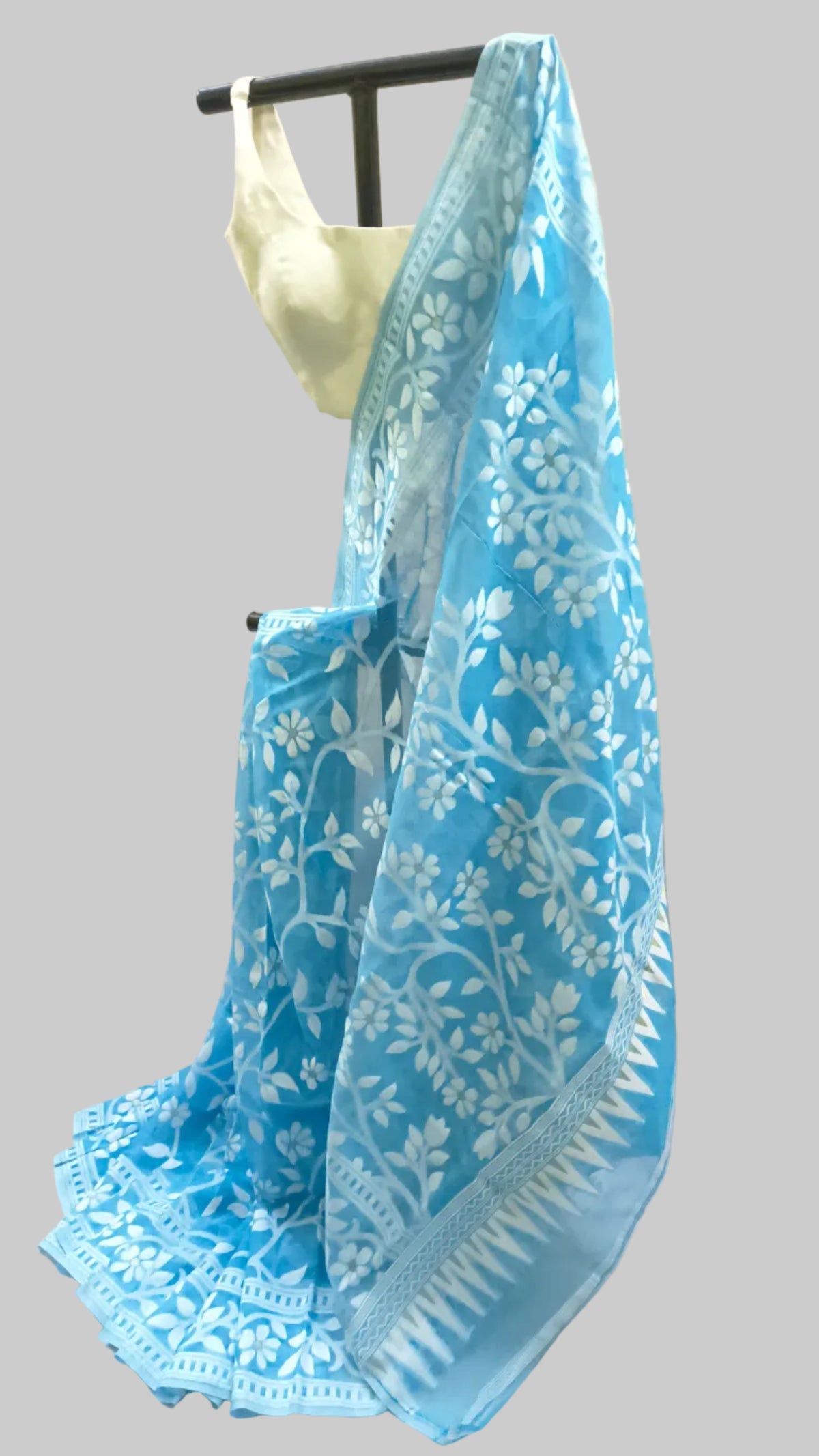 A light blue saree with white floral and geometric block-print patterns, draped over a black hanger with a cream-colored blouse visible at the top, set against a draped white fabric backdrop.