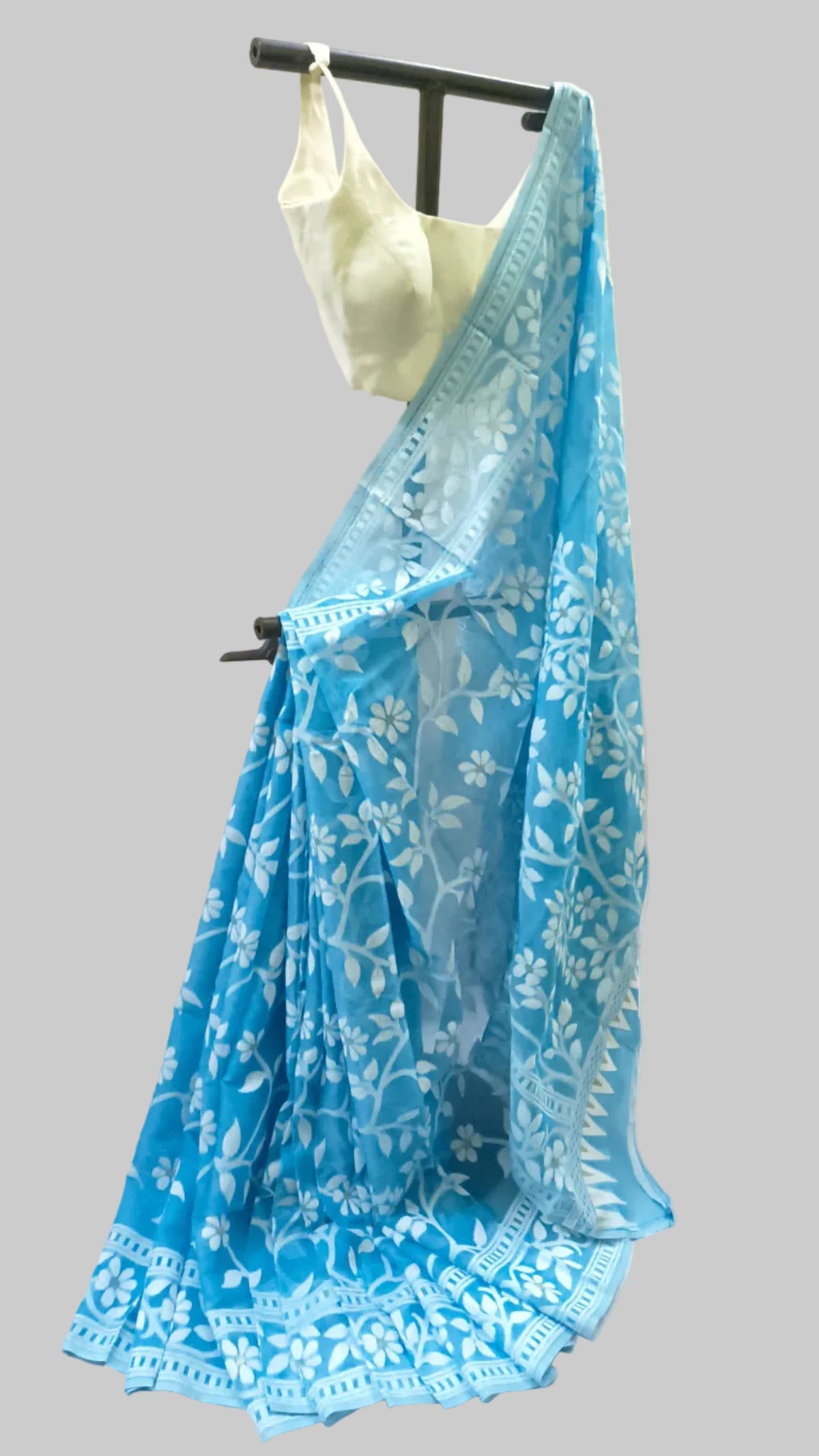 A light blue saree with white floral embroidery and a matching embroidered border, draped on a black hanger stand with a white blouse, set against a draped white fabric backdrop.