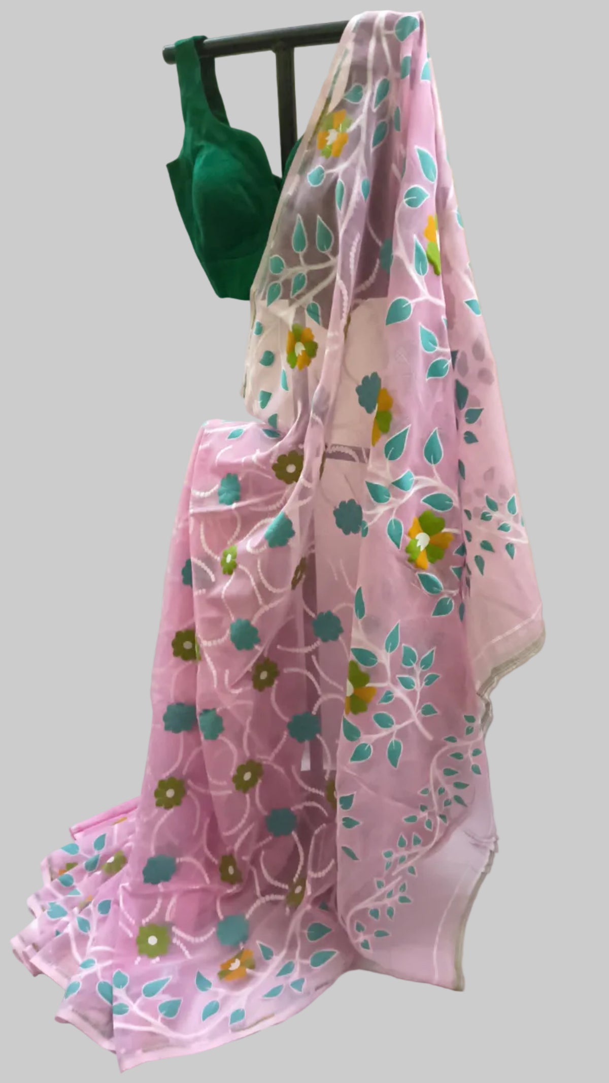 A pink organza saree with blue and yellow floral embroidery draped over a black hanger, with a dark green blouse visible at the top, set against a cream-colored fabric backdrop.