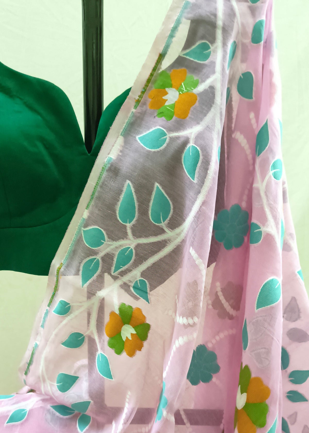 A pink saree with a sheer white border decorated with teal leaf patterns and orange and green floral motifs, draped over a dark green blouse.