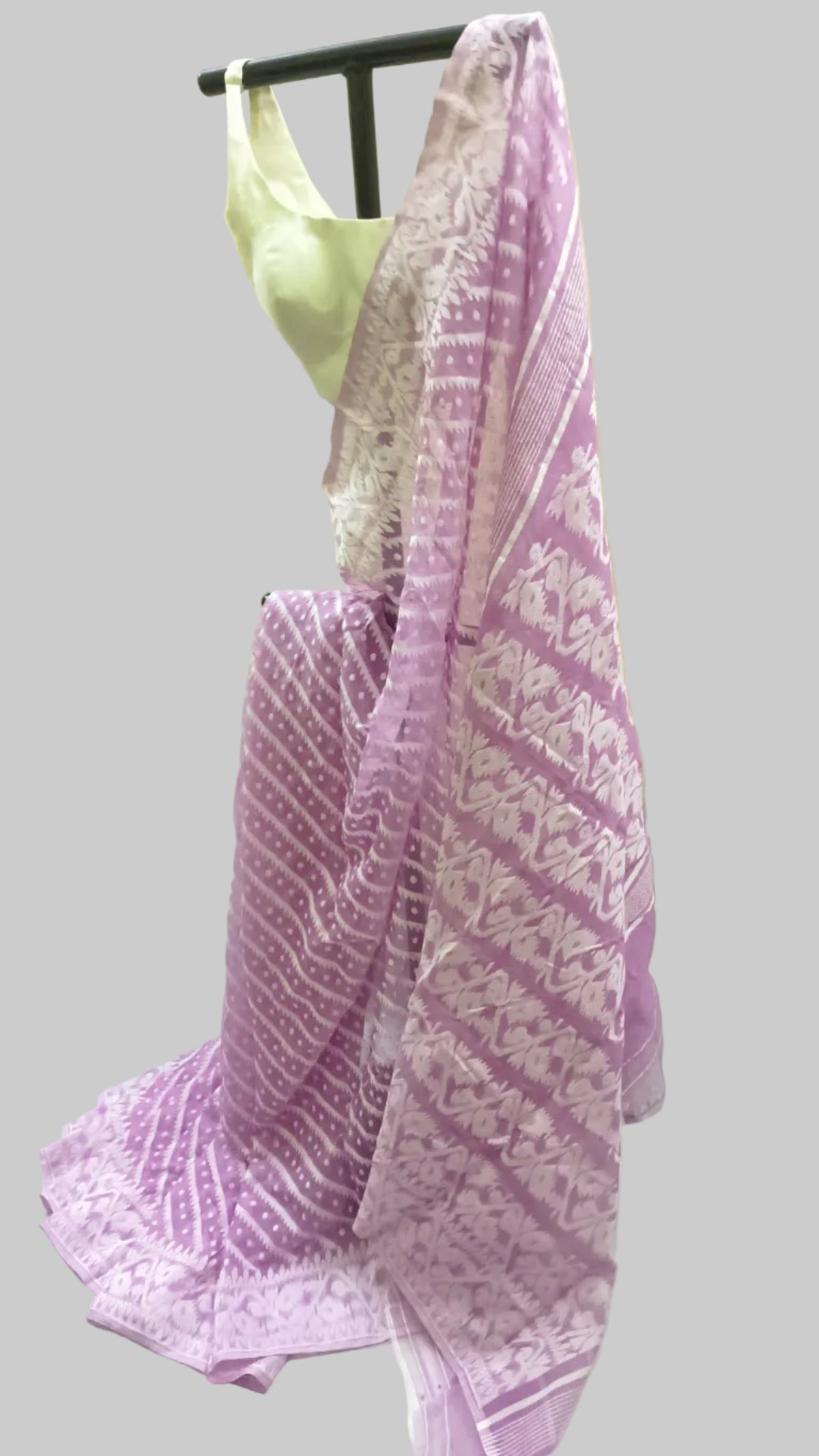A lavender and white patterned saree draped on a black dress form, with a light green blouse partially visible behind it, set against a cream-colored fabric backdrop.