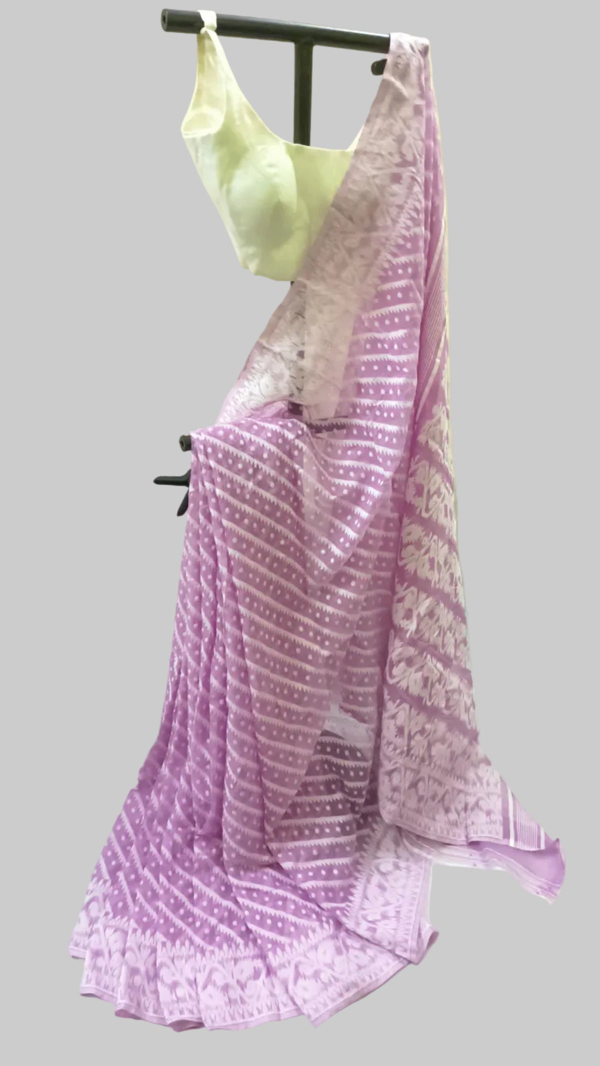 A lavender purple saree with white geometric patterns and a matching light green blouse, displayed on a black clothing hanger against a draped cream fabric backdrop.