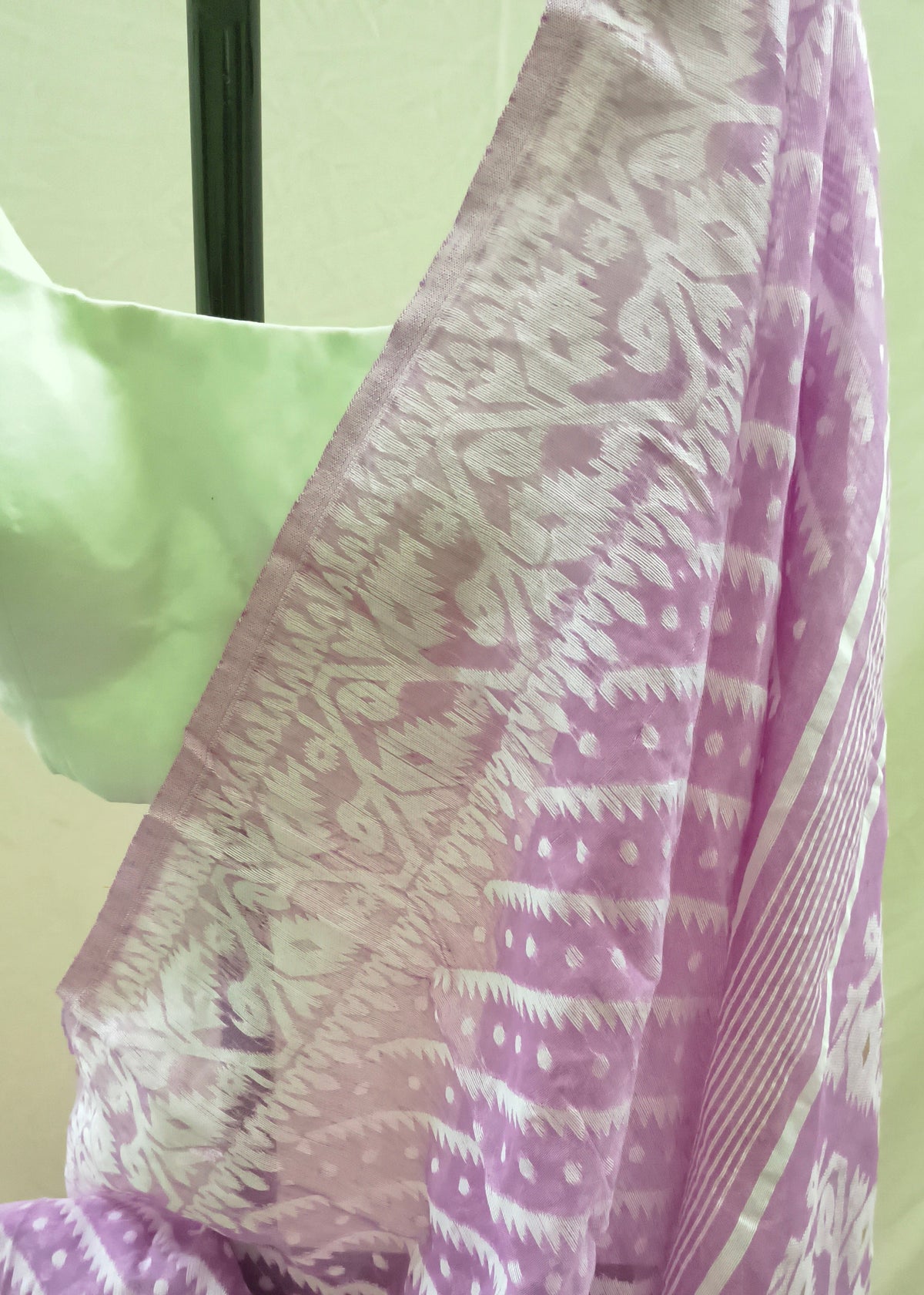 A close-up of a lavender and white patterned saree draped on a mannequin, showing the detailed ikat and tie-dye motifs alongside a light green blouse.