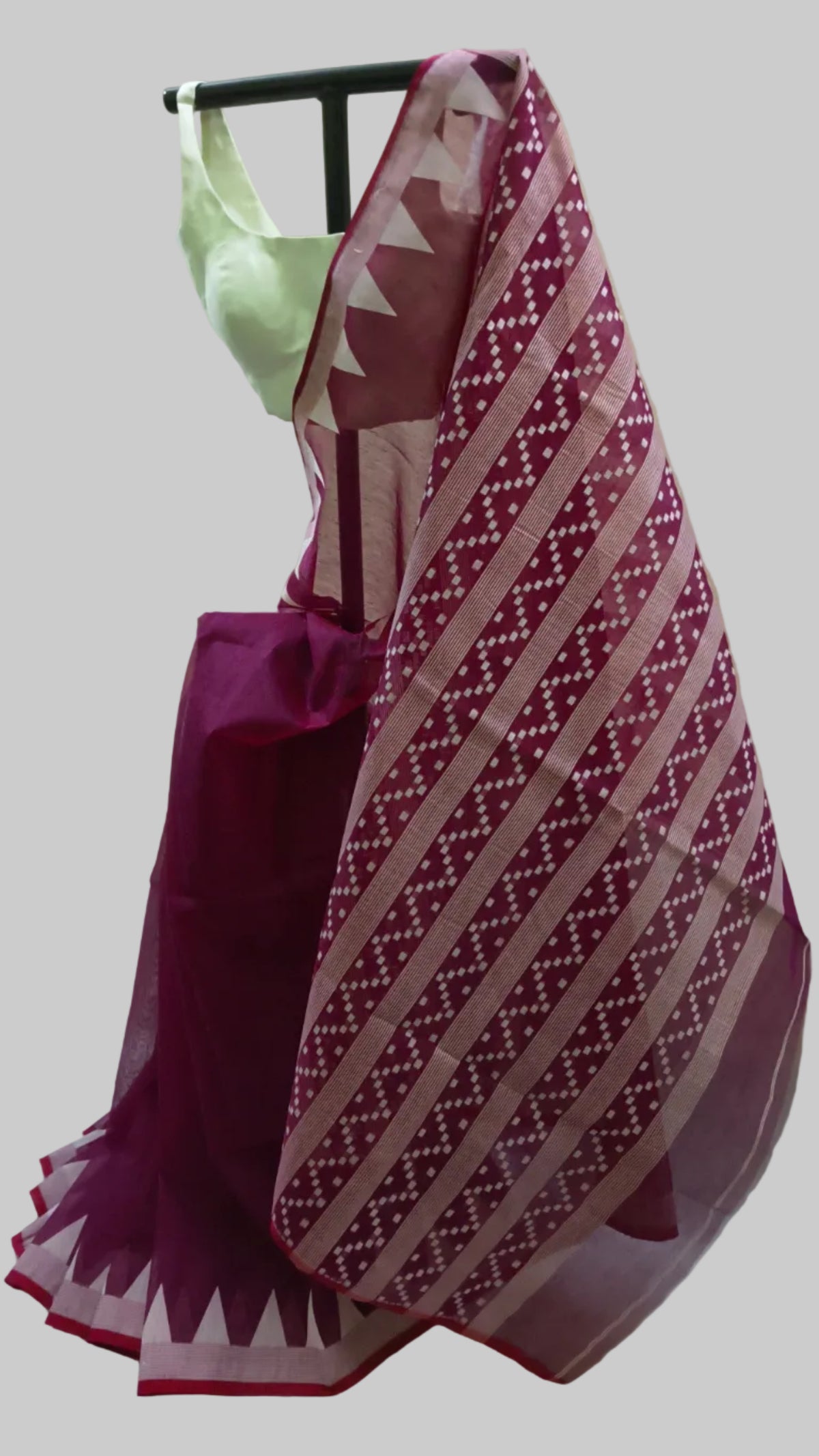 A traditional Indian saree displayed on a black hanger against a cream-colored fabric backdrop. The saree features a deep magenta-purple body with a wide border of white geometric patterns, and is paired with a light green blouse.