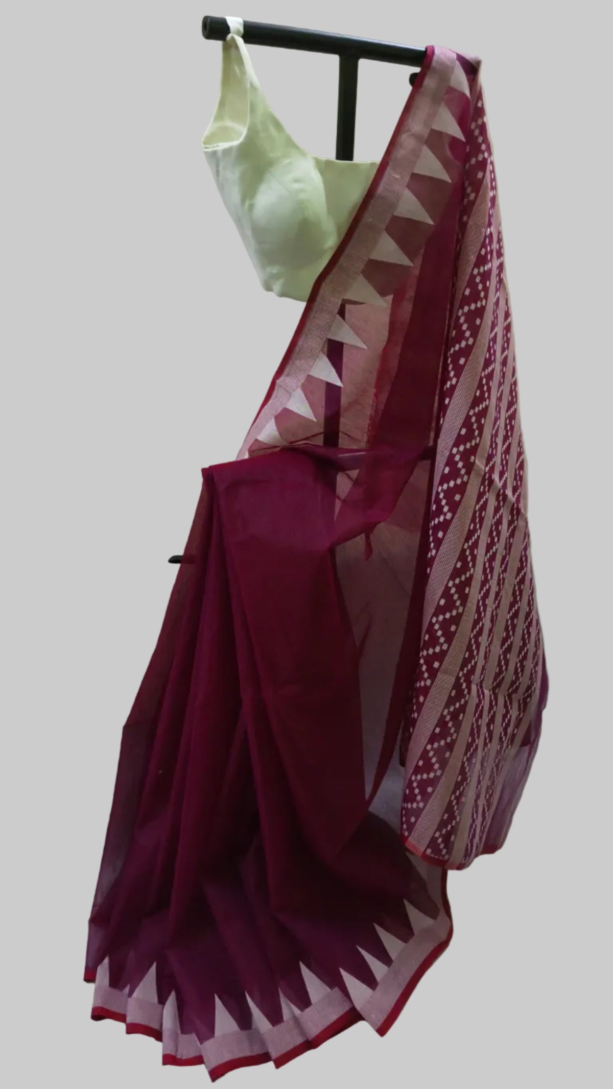 A deep magenta saree with a white and magenta geometric border pattern, draped on a black hanger with a white blouse, set against a draped white fabric backdrop.