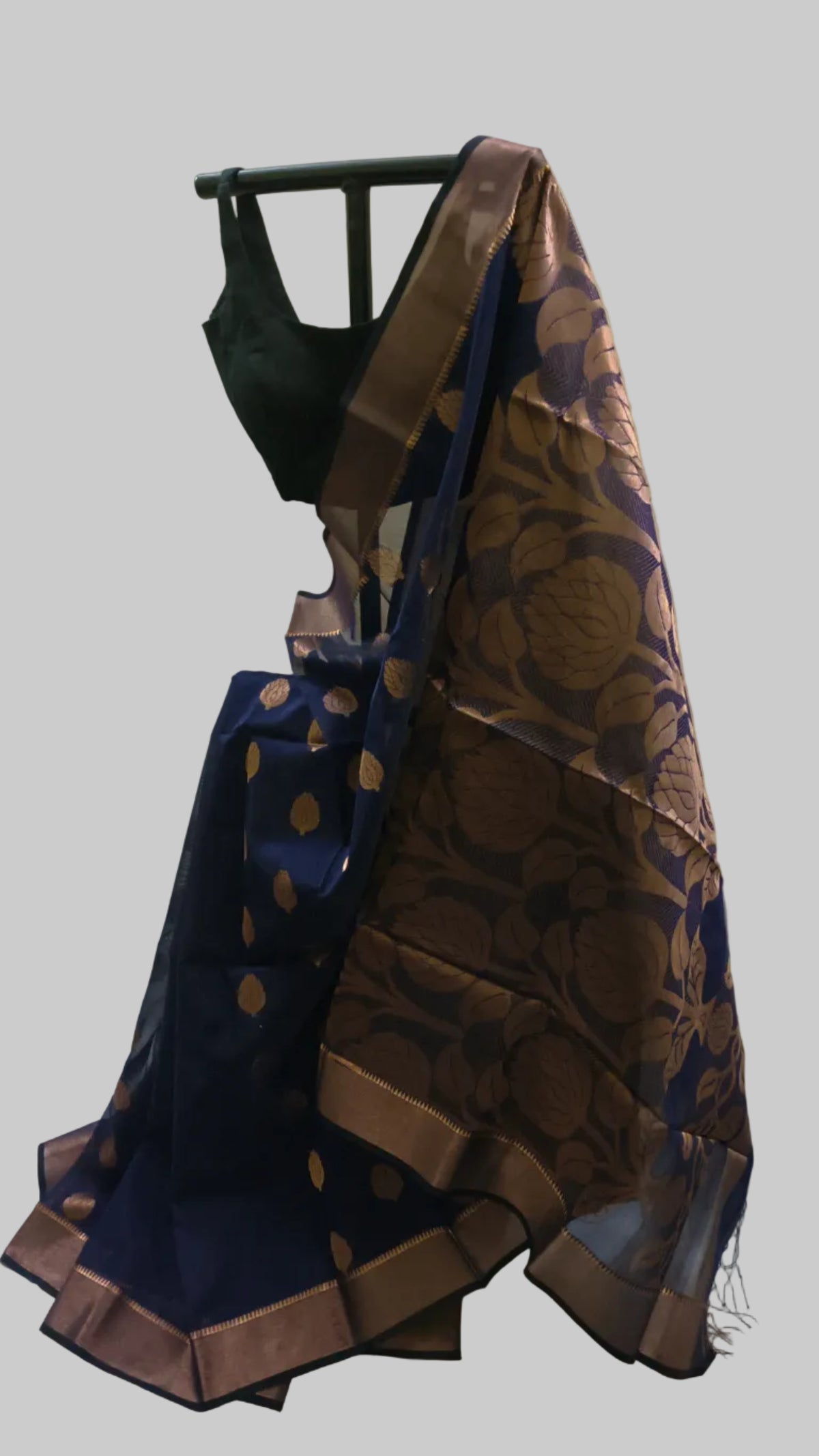 A dark blue and brown silk saree with gold zari floral motifs, draped on a black hanger with a black blouse, set against a cream-colored fabric backdrop.