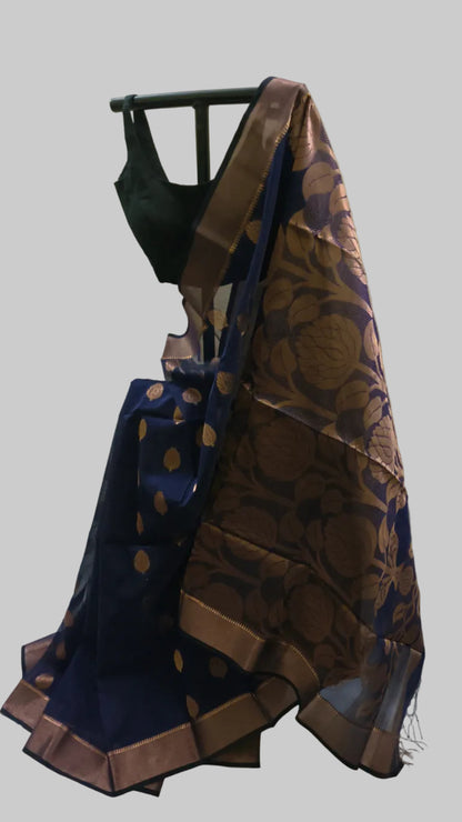 A dark blue and brown silk saree with gold zari floral motifs, draped on a black hanger with a black blouse, set against a cream-colored fabric backdrop.